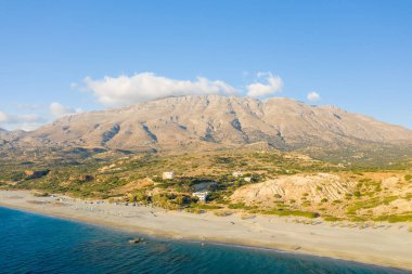 Yunanistan 'ın Girit kentindeki Triopetra plajının hava perspektifi, turkuaz deniz buluşması geniş kumlu sahil ve dramatik kayalık dağlarla açık mavi bir gökyüzünün altında yükseliyor..