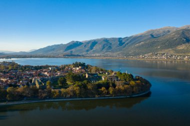 Geniş bir hava perspektifi, Ioannina Gölü ile çevrili tarihi Ioannina yarımadasını ve arka planda yükselen engebeli Pindus dağlarını yakalıyor. Sahnede kırmızı kiremitli çatılar, ortaçağ duvarları ve canlı bir suyun altında sakin bir su yer alıyor.