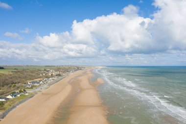 Omaha Sahili 'nin havadan görünüşü, yumuşak dalgalarla çevrili geniş kumlu bir kıyı şeridini ve parlak mavi gökyüzünü vuran kümülüs bulutlarıyla dolu bir kıyı evini gözler önüne seriyor. Sahne açık, taze ve davetkar..