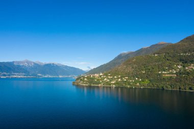 Cannobio 'nun geniş manzarası kuzey İtalya' daki Maggiore Gölü 'nün derin mavi sularının üzerindeki yeşil tepelere tünemişti. Manzarada dağınık evler, yemyeşil bitki örtüsü ve canlı, bulutsuz bir gökyüzünün altındaki uzak dağlık tepeler yer alıyor..