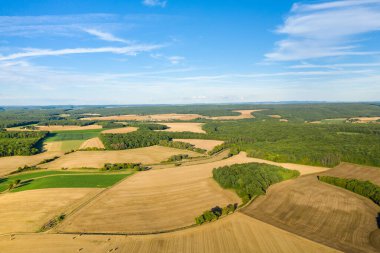 Açık mavi gökyüzünün altında, yoğun yeşil ormanlık alanla karışmış altın hasat edilmiş alanların havadan görünüşü. Geniş kırsal alan geometrik şekiller, doğal dokular ve yaz aylarında orta Fransa 'nın tipik açık kırsal bölgelerini yakalar..