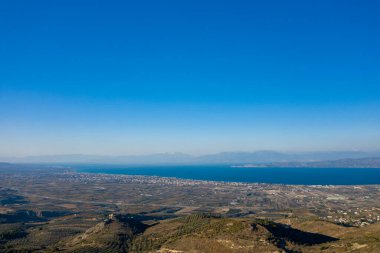 Uzak dağlar ve bulutsuz gökyüzü ile, derin mavi bir denize doğru uzanan, güneşli bir tarım vadisinin geniş bir görüntüsü. Manzara açık, huzurlu ve doğal ışıkla dolu..