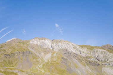 Yumuşak güneş ışığı, Gavarnie Gedre yakınlarındaki Pireneler 'de açık mavi bir gökyüzünün altında, üzerinde çimen parçaları olan yumuşak bir dağ tepesini vurguluyor. Manzara açıklık ve doğal sükuneti iletir..