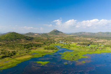 Hava manzarası, Kamboçya 'nın başkenti Kampong Chhnang' da Brateak Krola Gölü yakınlarında canlı bir mavi gökyüzü altında yavaşça kayan tepelere doğru uzanan yemyeşil sulak alanlar ve dağınık ağaçlar gösteriyor. Sudaki bitki örtüsü ve açık sular doğal desenler yaratır.