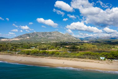 Bereketli yeşilliklerle çevrili sakin kumlu bir sahilin havadan perspektifi, arka planda yükselen engebeli dağlar, Orosei, Sardunya 'daki tüylü bulutlarla kaplı bir gökyüzünün altında..