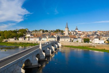La Charite sur Loire 'a uzanan tarihi taş köprünün üzerinde parlak bir gün ışığı. Sıra sıra geleneksel evler, önemli bir kilise kulesi ve Loire nehri boyunca yemyeşil bir alan. Sahne, ortaçağ mimarisinin ahenkli bir harmanını yakalıyor.