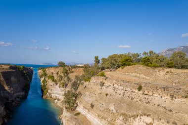 Korint Kanalı 'nın yüksek açılı görüntüsü. Dik, güneşli kireçtaşı uçurumlarını keserek seyrek bitki örtüsü ve mavi suyla birlikte berrak bir gökyüzünün altındaki uzak denize doğru açılıyor..