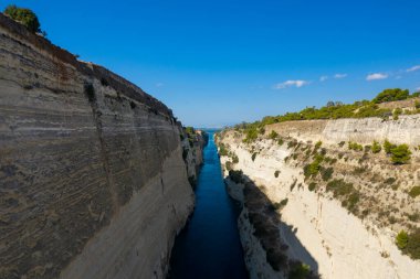 Uzun, derin su yolu parlak mavi bir gökyüzünün altındaki solgun kaya duvarlarını kesiyor. Dağınık yeşil çalılar ve uzak köprüler görülebiliyor. Güneş ışığı sahnesinde keskin jeolojik katmanlar ve çarpıcı doğal kontrastlar vurgulanıyor..