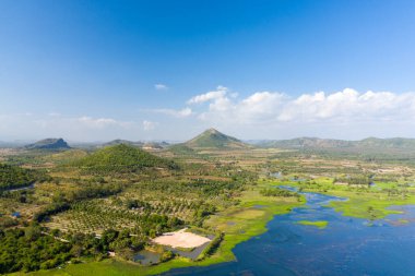 Hava perspektifi, Kamboçya 'nın Kampong Chhnang kentindeki Brateak Krola Gölü' nün mavi sularının etrafında dönen yeşil tepeler ve geniş sulak alanlar gösteriyor. Bitki örtüleri ve dağınık tarım arazileri uzak tepeleri çerçeveli, açık bir gökyüzünün altında uzanır.