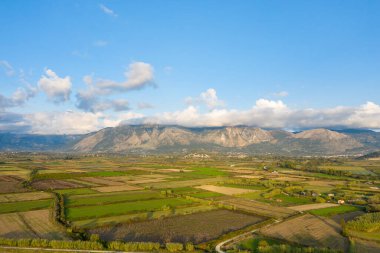 Uzakta engebeli dağlar ve dağınık bulutlarla geniş bir ovada ekilmiş tarlaların hava görüntüsü yama kalıbı oluşturuyor. Kırsal Yunan manzarası, geometrik şekiller ve doğal şekillerin vurgulandığı yumuşak öğleden sonra ışığıyla yıkanır.