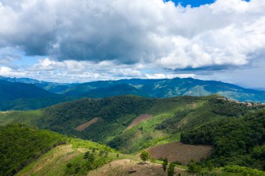 Yuvarlanan yeşil tepelerin ve yamalı tarım arazilerinin panoramik manzarası, Laos 'un Phou Khoun ve Luang Prabang kentlerinin kırsal kesimlerinde dramatik bulutların altında uzanır. Manzarada sık orman, ekili yamaçlar ve uzak mavi tepeler bulunur.