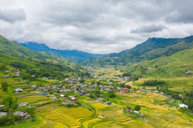 Teraslı pirinç tarlalarının geniş havası ve Vietnam yakınlarındaki Sapa yakınlarındaki yemyeşil vadide yuva yapmış dağınık köy evleri. Dramatik bulutlu gökyüzü ve katmanlı dağlar canlı bir kırsal manzara yaratır..