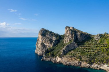 Dik kireçtaşı kayalıkları, derin mavi suların üzerinde yükselir, antik bir taş kalıntısı tarafından taçlandırılır ve teraslı yeşilliklerle çevrilidir. Açık gökyüzü ve canlı kıyı manzarası İtalyan kırsalının sakin atmosferini çağrıştırıyor..