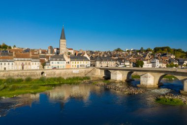Loire Nehri boyunca Pastel Evlere ve La Charite sur Loire 'ın uzun manastır kulesine uzanan tarihi taş köprü kemerleri. Parlak güneş ışığı ve parlak mavi gökyüzü Loire Vadisi 'ndeki ortaçağ mimarisi ve nehir kenarı cazibesini ön plana çıkarıyor..