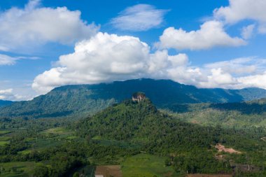 Geniş kırsal bir vadide yemyeşil bir ormanın üzerinde, uzak dağların ve dalgalı bulutların zeminine kurulmuş, tek bir düzlük tepe yükselir. Sahne, Güneydoğu Asya 'nın katmanlı bitki örtüsü ve doğal dokularını vurguluyor.