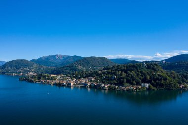 Kuzey İtalya 'daki Orta Gölü boyunca yoğun ormanlık tepelerin eteklerine kurulmuş göl kenti manzarası. Sakin mavi su, çevresini çevreleyen yeşilliği ve berrak gökyüzünün altındaki uzak dağ yamaçlarını yansıtır..