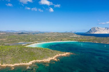 San Teodoro, Sardinya yakınlarındaki Cala Brandinchi sahilinin hava perspektifi, geniş kıvrımlı bir körfez, canlı turkuaz su, demirlemiş tekneler ve açık mavi bir gökyüzü altında etkileyici bir dağ altında yemyeşil bir manzara..