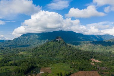 Yoğun tropikal orman ve yama tarım arazilerinin üzerinde, gökyüzünün altında geniş ve dalgalı bulutlarla dolu geniş bir dağ zeminine kurulmuş, belirgin düz bir tepe yükseliyor. Manzara doğal dokuları ve katmanlı yeşilliği vurguluyor.
