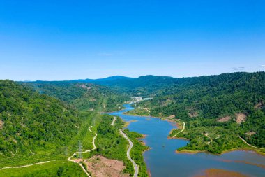 Hava perspektifi, yoğun ormanlık tepeler ve engebeli arazi arasında canlı mavi bir gökyüzü altında kıvrımlı bir nehir olduğunu ortaya koyuyor. Manzara dolambaçlı yollar, yemyeşil bitki örtüsü ve uzak bir doğal güzellik hissine sahiptir..