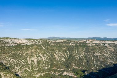 Navacelles Sirki 'ndeki canlı mavi gökyüzünün altında yoğun bitkisel yamaçların üzerinde dev kireçtaşı kayalıklar yükselir. Engebeli dokular ve katmanlı kaya oluşumları parlak güneş ışığı ile vurgulanarak doğal bir ihtişam hissi yaratıyor..