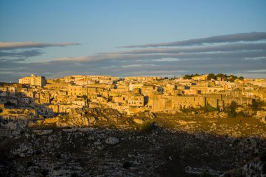 Matera 'nın antik taş binaları sıcak öğleden sonra ışıklarıyla aydınlanır. Önünde dramatik kayalık bir arazi ve yukarıda katmanlı bulutlarla kaplı bir gökyüzü vardır..