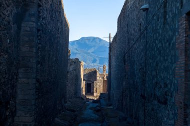 İtalya 'nın Pompeii kentinde, yüksek volkanik taş duvarlarla çevrili dar bir ara sokak, güneşli harabelere ve berrak mavi gökyüzünün altındaki uzak bir dağa doğru açılıyor. Sahnede derin gölgeler, pürüzlü duvarcılık ve ikonik arkeolojik manzara yer alıyor..