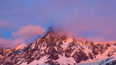 Canlı pembe ve turuncu bulutlar şafak vakti Aiguille du Midi 'nin engebeli, karla kaplı tepesinde sürükleniyor, dramatik kaya dokuları ve yumuşak ışık ile Alp manzarasını aydınlatıyor..