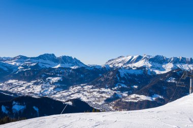 Karlı bir kayak yamacında parlak bir kış günü. Megeve köyünün panoramik manzarası, dramatik alp zirveleri ve derin mavi gökyüzü canlı, canlandırıcı bir atmosfer yaratıyor..