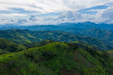 Parlak güneş ışığı yeşil tepeleri ve dağınık ağaçları vurguluyor, Laos 'un Phonsavan ve Phou Khoun arasındaki kırsal bölgede dramatik bulutlarla dolu gökyüzünün altında uzanan uzak mavi tepeler. Sahnede yemyeşil bitki örtüsü var.