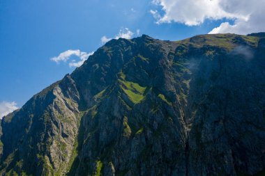 Parlak güneş ışığı altında yeşil çimen parçalarıyla sarp engebeli kaya yüzeyleri ve Pireneler dağlarında dağınık bulutlar. Derin gölgeler, çentikli dokuları ve manzaranın cesur hatlarını vurgular..