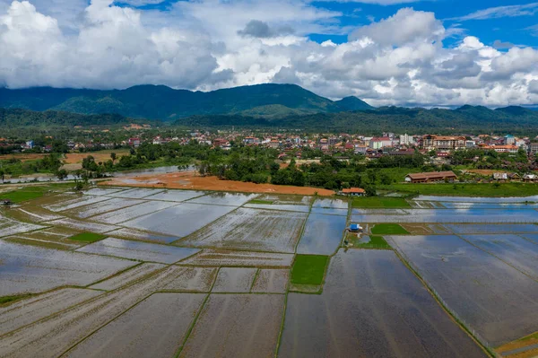 Yansıtıcı pirinç tarlaları, yemyeşil ve binalarla çevrili Vang Vieng, Laos 'un kenarına doğru uzanıyor. Uzak orman dağlarının üzerinde asılı dramatik bulutlar, yağmur mevsiminde kırsal araziyi vurguluyor..