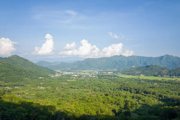 Dağılmış köyleri ve sık ormanları olan yemyeşil bir vadinin panoramik görüntüsü, açık mavi gökyüzünün altında dramatik kümülüs bulutları olan yuvarlanan dağlar tarafından çerçevelenmiş. Manzara geniş, güneşli ve huzurlu görünüyor..