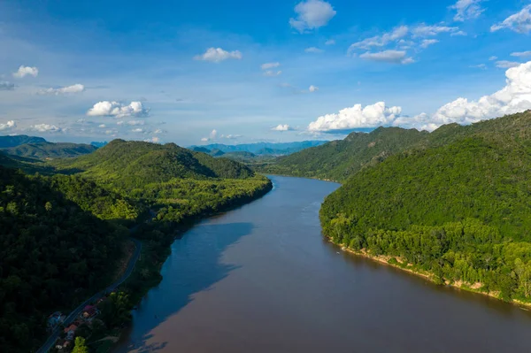 Mekong Nehri 'nin geniş bir hava perspektifi, canlı mavi gökyüzünün altında, gür, ormanlık tepeler ve dağınık bulutlar arasında süzülüyor. Manzarada canlı yeşillikler, yuvarlanan arazi ve Güneydoğu Asya 'daki uzak sıradağlar yer alıyor..