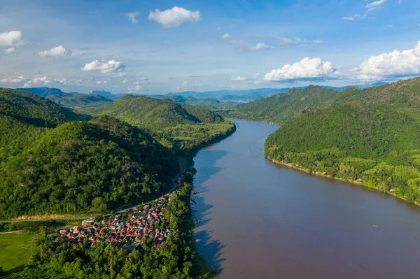Sarmal Mekong nehri boyunca uzanan yemyeşil tepelerin tabanında yuva yapmış küçük bir nehir kenarı köyünün geniş hava manzarası. Manzarada yoğun tropikal bitki örtüsü, kırmızı çatılar ve Güneydoğu 'da parlak mavi bir gökyüzü altında geniş bir doğal manzara yer alıyor.