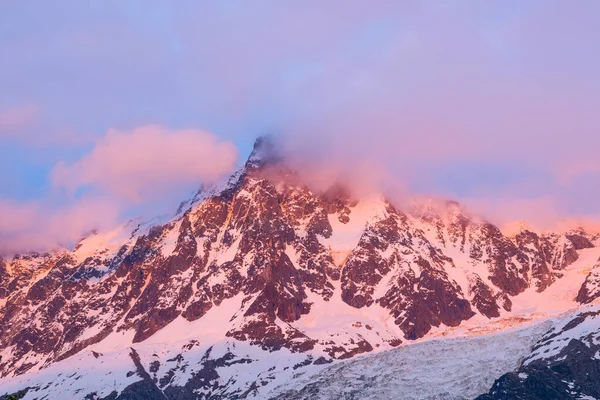 Yumuşak pembe ve lavanta bulutları, güneş doğarken Aiguille du Midi 'nin engebeli, tozlu zirvesinde Fransız Alpleri' nin dramatik dağ yüzeyine hafif bir ışık saçıyor..