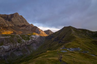 Altın akşam güneşi, Fransız Pireneleri 'nde dramatik çimenli bir tepe ve kayalık uçurumlar üzerinde otluyor. Kasvetli bir gökyüzünün altında küçük bir park alanı bulunuyor. Manzara cesur dokular, katmanlı yamaçlar ve sakin bir atmosfere sahiptir..
