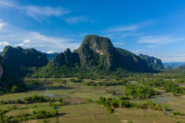 Dramatik karst dağları yeşil tarlaların ve Laos kırsalındaki dağınık su havuzlarının üzerinde yükseliyor. Açık arazide canlı mavi gökyüzü, yemyeşil bitki örtüsü ve Güneydoğu Asya 'nın tipik doğal desenleri yer alıyor..