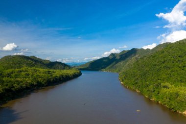 Mekong nehri yoğun ormanlık tepeler arasında savruluyor. Canlı mavi bir gökyüzü altında, dağınık bulutlar ve uzak dağ sırtları ile. Güneş ışığı nehir kıyıları boyunca uzanan zengin yeşil bitki örtüsünü vurguluyor, dingin ve geniş bir manzara yaratıyor..