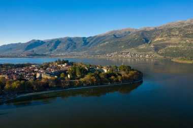 Geniş bir hava perspektifi, Ioannina Gölü ile çevrili tarihi eski Ioannina kasabasını ve arka planda yükselen Pindus dağlarını görüntüler. Sahnede kırmızı kiremitli çatılar, ortaçağ duvarları ve canlı mavi gökyüzünün altında sakin su yer alıyor.