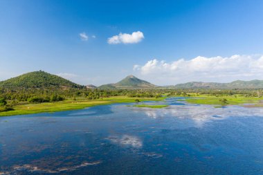 Brateak Gölü 'nün canlı mavi suları Kamboçya' nın Kampong Chhnang şehrinde açık bir gökyüzü altında yuvarlanan yeşil tepelere ve dağınık ağaçlara doğru uzanıyor. Su bitkileri ve açık otlaklar kıyı şeridi boyunca doğal desenler oluşturur..