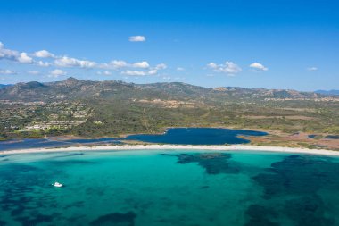 Sardunya 'daki Spiaggia Lu Impostu' nun havadan görünüşü turkuaz suyu, beyaz kumlu bir kıyı şeridi ve yeşil tepeler ve dağınık bulutlar üzerine kurulmuş koyu mavi bir lagün gösteriyor..