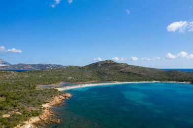 Geniş bir hava manzarası Spiaggia la Salinedda, Sardinya 'da hilal şeklinde bir körfez olduğunu gösteriyor. Bereketli yeşil tepeler, kayalık kıyı şeritleri ve dağınık bulutlarla dolu açık mavi bir gökyüzünün altında..