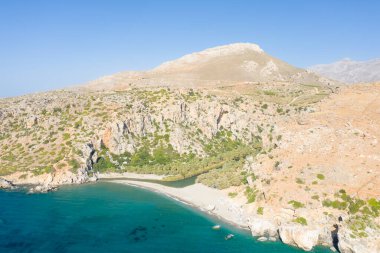 Dar bir ırmağın, engebeli kayalık tepelerle çevrili turkuaz denizle buluştuğu ve Girit 'teki berrak mavi gökyüzünün altında seyrek yeşillik olan sessiz kumlu bir körfezin havadan görünüşü..
