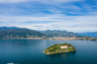Isola Madre 'nin gökyüzü perspektifi Maggiore Gölü' nün derin mavi suları ile çevrili ve ön planda tarihi bir villa ve yemyeşil bahçeler bulunmaktadır. Arka planda göl kenti, ormanlık tepeler ve gökyüzünün altındaki dağlık tepeler görünüyor.