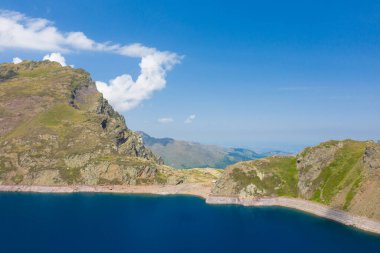 Pireneler 'deki Lac Bleu' nun kıtır yaz manzarası, engebeli yeşil yamaçlar ve kayalık uçurumlar parlak bulutlarla dolu, mavi bir gökyüzünün altında yükseliyor..