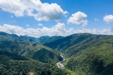 Dolambaçlı bir yolu olan yemyeşil bir vadinin geniş manzarası ve canlı mavi bir gökyüzünün altında dağılmış kırsal binalar ve Phou Khoun ile Luang Prabang, Laos arasındaki yolda tüylü bulutlar. Manzara katmanlı