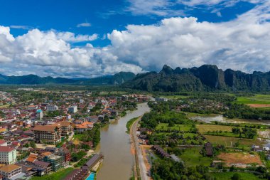 Vang Vieng, Laos 'un yüksek açılı manzarası, Vietnam Song Nehri kasabada kıvrılıyor ve arka planda dramatik kireçtaşı kayalıkları yükseliyor. Sahnede yamalı pirinç tarlaları, dağınık binalar ve canlı mavi gökyüzünün altında yemyeşil alanlar yer alıyor.