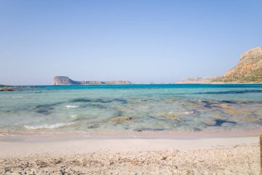 Şeffaf sığ sular, bulutsuz mavi gökyüzünün altında Girit 'in Balos şehrinde kayalık çıkıntıları olan kumlu bir kumsalla buluşur. Sahne güneşli, sakin ve doğal olarak enerjik..