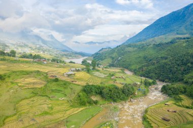 Vietnam, Sapa 'nın yemyeşil vadisinde katmanlı pirinç terasları ve dolambaçlı bir nehir manzarası. Alçak bulutlar uzak dağlarda sürükleniyor, sakin ve atmosferik kırsal bir manzara yaratıyor..