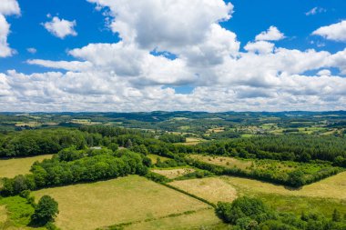 Yama alanları, sık yeşil ormanlık alanlar ve kabarık beyaz bulutlarla dolu gökyüzünün altındaki kayan tepelerin yer aldığı güneşli kırsal alan manzarası. Manzara bir açıklık ve doğal sükunet hissi yayar..